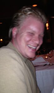 Jason,  sits at a  dining table wearing a neutral colored shirt. His blond hair and bright smile is caught strikingly in the warm candle lit atmosphere.
