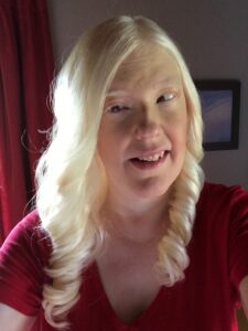 Photo description of Chrys, who is wearing a red top. Her hair is long, curly, and platinum blonde. She is smiling and looking slightly to her left. They are indoors, with a deep red curtain and part of a framed picture visible in the background. The lighting highlights her hair and face, casting some shadows on the right side. 