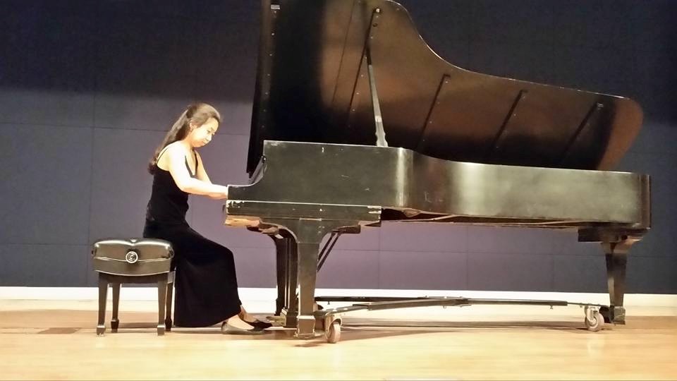 Natalie is dressed in a formal black dress playing a grand piano on a wooden stage. She is seated on a piano bench, focused on the keys as she plays. The background is a dark, solid color wall, suggesting a performance or practice space. The grand piano has its lid open, with visible strings and internal components. The lighting highlights the pianist and piano, while the rest of the scene remains simple and uncluttered.