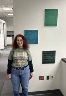 Laura Rae stands in front of a white wall decorated with several square pieces of artwork. The panels are in shades of green and teal. She has long dark hair and is wearing a black top with blue jeans.